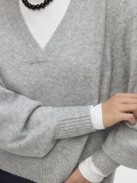 Grå stickad tröja med V-ringning, ribbade muddar och mjuk textur, lager-på-lager över en vit långärmad skjorta, som visar en avslappnad stil.
