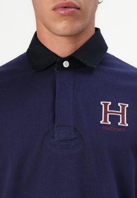 Polo de marinha com gola preta, textura de tecido suave, apresenta o logótipo bordado "H" em vermelho e branco, com frente abotoada e detalhes de costura visíveis.