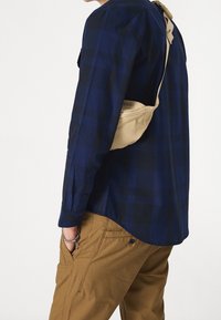 Beige axelväska som bärs över en skjorta i marinblå och svart rutigt mönster med långa ärmar, ihop med beige byxor. Har kontrasterande färger och texturer.