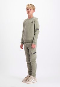 Olijfgroene sweatshirt en joggers van zacht materiaal, met een klein logo op de borst, cargozakken op de broek en neutrale sneakers.