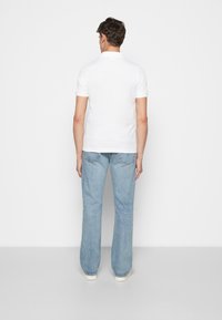 Polo blanc à manches courtes, coutures visibles, personne debout portant un jean bleu évasé, chaussures de couleur claire, fond uni.