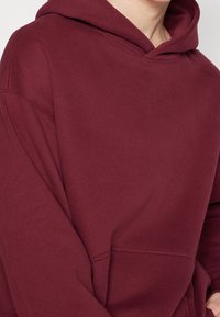 Sudadera burdeos con capucha, que presenta un bolsillo tipo canguro, textura suave y detalles de costura, diseñada para el uso casual.