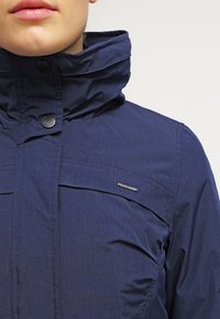 Giacca blu navy con colletto alto, bottoni a scatto e tessuto a trama; caratterizzata da un logo sul petto e tasche orizzontali.