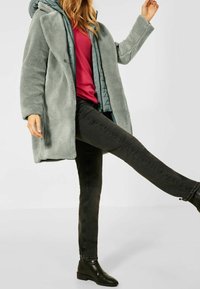 Faux fur-kappa i ljusgrön, lager på lager över en quiltad teal-jacka, bärs med en röd topp och svarta skinny jeans, matchat med svarta ankelstövlar.