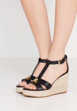 Sandales compensées en cuir noir avec un design à lanière T, des accents en métal doré et des talons compensés en jute. Fini lisse et orteils ouverts.