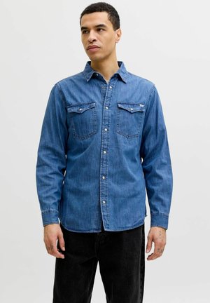 Mann trägt ein blaues Jeanshemd mit Knöpfen und zwei Brusttaschen sowie schwarze Hosen, steht vor einem schlichten hellen Hintergrund.