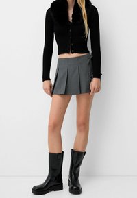 Svart rippad cardigan med fuskpälskrage, grå plisserad minikjol och knähöga svarta glänsande stövlar. Enkelt design med minimala detaljer.