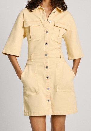 Vrouw draagt een lichtgele jurk met knoopsluiting, halflange mouwen en meerdere zakken, handen nonchalant in de zijzakken.