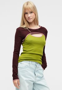 Μπλούζα μακρυμάνικη με σχέδιο κοψίματος. Σκούρα καφέ μανίκια, πράσινο σώμα με ροζ περίγραμμα. Φορεμένη με ανοιχτό μπλε τζιν παντελόνι.