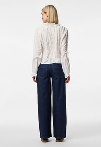 Blusa branca com detalhe de costas franzidas, mangas longas e gola alta, combinada com jeans de perna larga azul escuro e calçado bege.