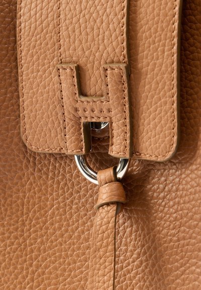 Κοντινό πλάνο δερμάτινης τσάντας με υφή από μικρές ρυτίδες σε ανοιχτό καφέ χρώμα, με ραμμένο λουρί που περνά μέσα από ασημένιο μεταλλικό κρίκο και στερεώνεται με δερμάτινο βρόγχο.