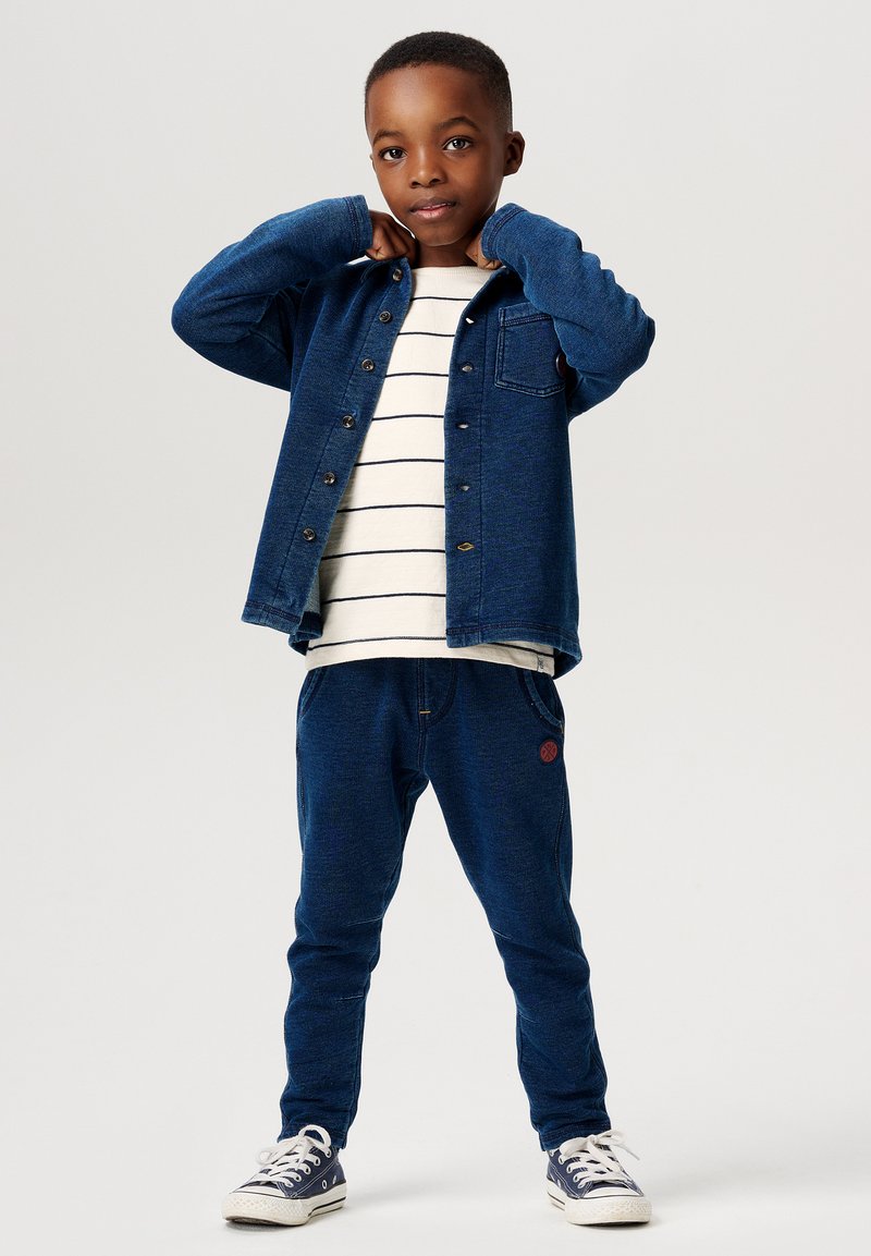 Blå denimskjorta med knäppning, vit randig t-shirt under, matchande blå sweatpants och marinblå sneakers med vita snören.