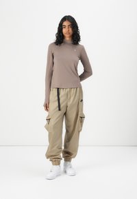 Langärmliges braunes Oberteil mit hohem Kragen, kombiniert mit beigen Cargo-Hosen mit Reißverschlusstaschen und elastischen Bündchen, abgerundet mit weißen Sneakers.