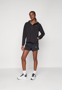 Svart lett jacket med hette, sammen med svarte shorts og hvite treningssko. Merkelig logo på jakken og shortsene.