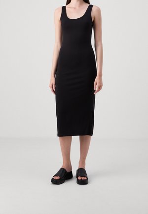 Kobieta w czarnej, bezrękawnikowej sukience midi i czarnych, platformowych sandałach na otwartym palcu stoi na jednolitym, jasnoszarym tle.