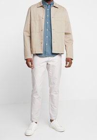 Veste en toile beige avec fermeture à boutons et deux poches avant, portée sur une chemise chambray bleue. Associée à un pantalon clair et des baskets blanches.