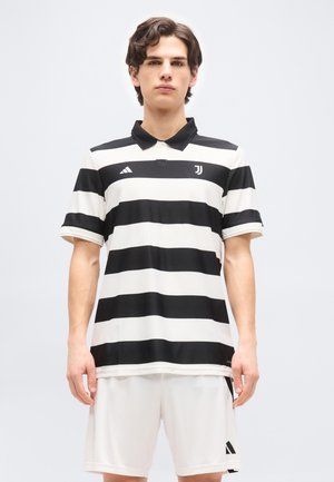 Junger Mann trägt ein schwarz-weiß gestreiftes Juventus-Fußballtrikot und weiße Shorts, steht vor einem schlichten hellen Hintergrund.