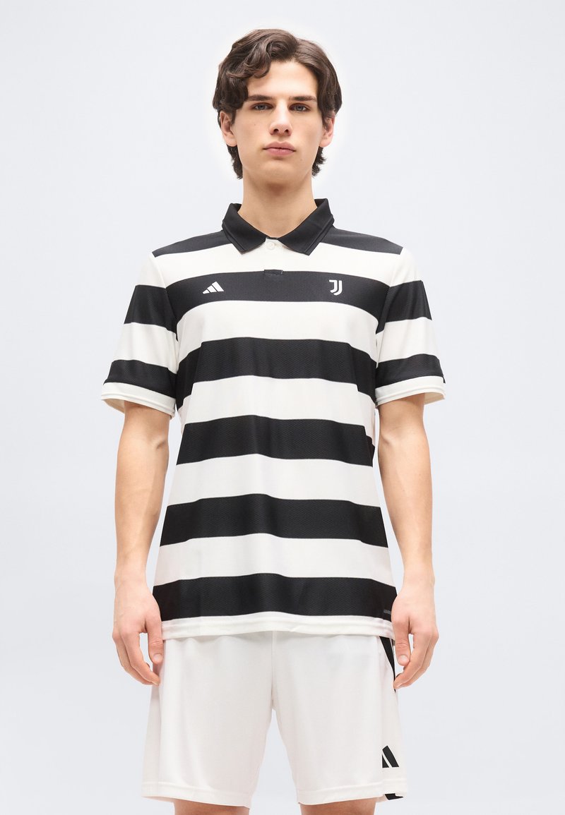 Νεαρός άνδρας φορώντας τη φανέλα ποδοσφαίρου της Juventus με μαύρες και άσπρες ρίγες και άσπρο σορτς, στέκεται μπροστά σε ένα απλό φωτεινό φόντο.
