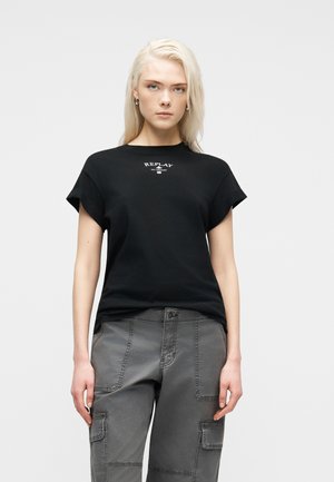 Νεαρή γυναίκα με μακριά ξανθά μαλλιά φοράει μαύρο t-shirt Replay και γκρι παντελόνι cargo, στέκεται μπροστά σε ένα απλό ανοιχτό γκρι φόντο.