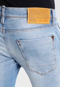 Ljusblå denimjeans med en tan-läderetikett med perforeringar vid midjan, kontrasterande sömmar och en vanlig bakfickdesign.