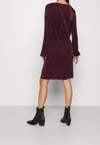 Burgunderfarbens, geripptes Kleid mit langenÄrmeln und tailliertem Schnitt, kombiniert mit schwarzen Stiefeletten mit einem klobigen Absatz. Glatte Textur.