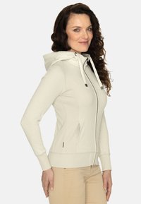 Felpa leggera beige chiaro in tessuto morbido e testurizzato. Presenta una zip completa, cappuccio regolabile con cordino e tasche laterali. Vestibilità slim con polsini a costine.