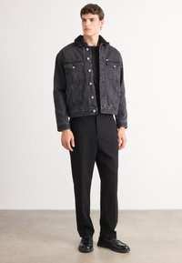 Chaqueta de mezclilla negra con cuello de borrego, cierre frontal de botones, dos bolsillos en el pecho, combinada con pantalones y zapatos negros, sobre un fondo liso.