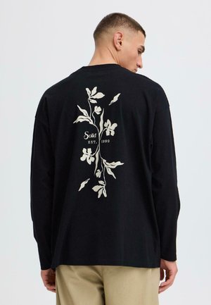 Homme portant un t-shirt noir à manches longues avec un motif floral blanc et le texte « Solid EST. 1999 » dans le dos, associé à un pantalon beige.