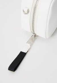 Borsa bianca dalla texture morbida con una zip metallica, caratterizzata da una tracolla nera e dettagli in argento sul tirante della zip.