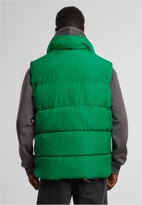 Grön vadderad väst med hög krage, sydda sektioner och en jämn struktur, bäddad över en grå långärmad topp.