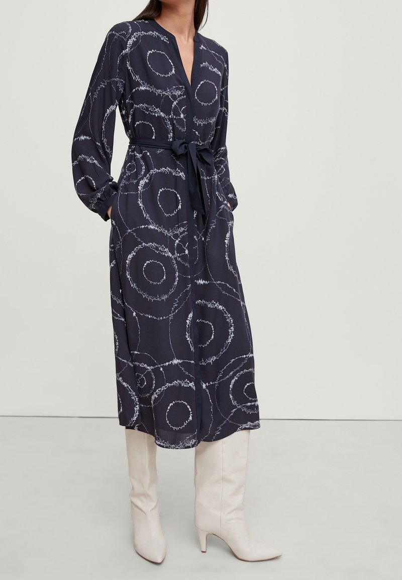 Abito navy con motivi circolari bianchi, maniche lunghe, scollatura a V e cintura. Indossato con stivali crema fino al ginocchio. Tessuto liscio e design fluido.