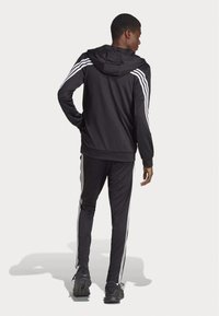 Ensemble de sport noir comprenant une veste à capuche et un pantalon ajusté, avec des accents à trois stripes blanches le long des manches et des jambes.
