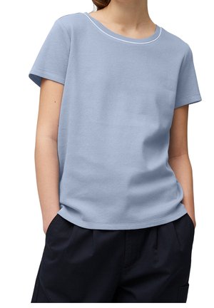 Persoon die een lichtblauw gestructureerd kortemouwenhemd en donkere losse broek draagt met handen in de zakken, op een witte achtergrond.