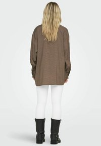 Brązowa koszula w kratę o oversize'owym kroju z długimi rękawami, połączona z białymi legginsami i czarnymi botkami za kostkę. Gładki materiał o luźnym kroju.