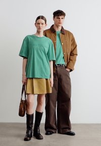 Η γυναίκα φοράει πράσινο πουκάμισο σε υπερμεγέθη γραμμή, κίτρινη καρό φούστα, μαύρες μπότες και κρατά καφέ τσάντα· ο άντρας φοράει καφέ τζάκετ, πράσινο πουκάμισο και καφέ παντελόνι.