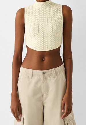 Haut court sans manches en maille crème avec motif torsadé, associé à un pantalon cargo beige porté par une personne sur un fond uni.