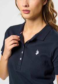 Kvinne som justerer kragen på en marineblå ribbestrikket poloshirt med tre knapper og en hvit logo av en polo-spiller på brystet.
