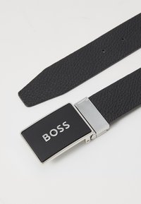 Schwarzlederner Gürtel mit strukturiertem Finish, ausgestattet mit einer rechteckigen silbernen Schnalle mit schwarzer Oberfläche, die mit "BOSS" graviert ist.