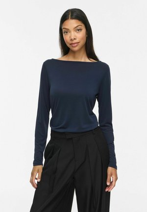 Jeune femme aux longs cheveux foncés porte un haut bleu marine à manches longues et col bateau et un pantalon noir plissé taille haute sur fond blanc.
