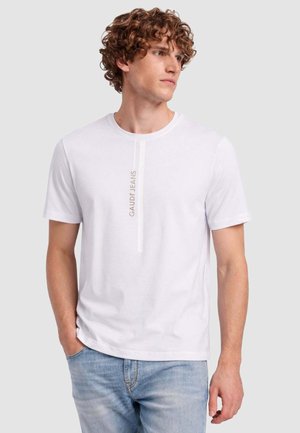 Giovane uomo con capelli ricci che indossa una maglietta bianca a maniche corte con la scritta verticale "GAUDÍ JEANS" e jeans azzurri, che guarda verso destra.