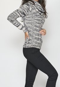 Pull tricoté rayé gris et noir avec col bénitier, manches ajustées et texture douce, associé à un jean noir avec des accents décoratifs.