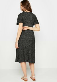 Robe noire à pois blancs ; manches courtes, mi-longue, design fluide. Des sandales plates avec des lanières légères complètent le look.