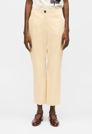 Beżowe, krótko przycięte spodnie o prostych nogawkach z czarnym guzikiem, noszone z brązowymi butami typu Mary Jane i jasnym t-shirtem z nadrukiem.