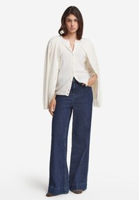 Vit knapp-up kofta med en avslappnad passform över en ljus topp och mörka vida jeans. Modellen står med ena handen i fickan, neutral bakgrund.