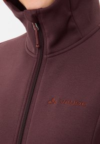 Bordeaux fleecejack met hoge kraag, rits aan de voorkant en gestructureerde stof. Heeft een subtiele logo in contrasterend oranjebruin.