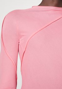 Gros plan sur une personne portant un Haut moulant à manches longues rose avec des détails de coutures visibles le long de l'épaule et du bras.