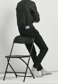 Svarta denimjeans och en mörk långärmad skjorta, sittande på en svart hopfällbar stol. Barfota för synligt, med minimal bakgrund.