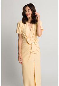 Sukienka sięgająca do kolan w żółtym kolorze, wykonana z miękkiego materiału, z krótkimi bufkami, głębokim dekoltem w kształcie litery V, rozcięciem z boku oraz kokardą w pasie.