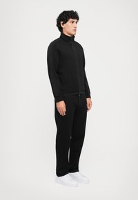 Homme debout contre un fond blanc, portant une veste zippée noire et un pantalon noir avec des baskets blanches, tourné sur le côté avec les mains détendues.