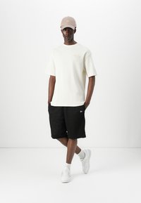 Cream ribbed short-sleeve t-shirt, black mesh shorts, beige cap, and white sneakers. Notable details include script logo on shirt and shorts.